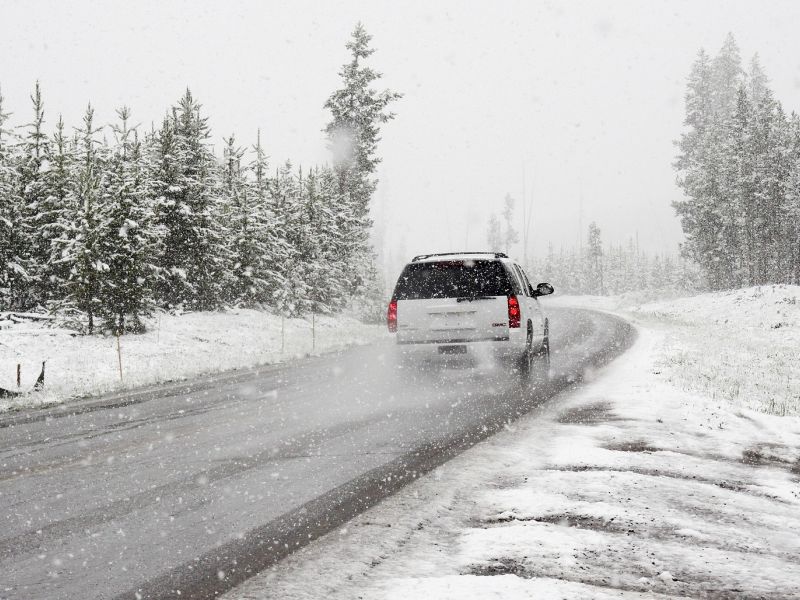 Na zdjęciu zaśnieżona droga, opady śniegu i sunący po drodze samochód rozbryzgujący kołami śnieg
